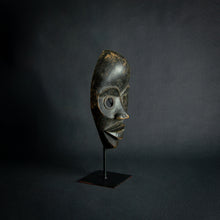 Load image into Gallery viewer, an mask from West Africa featuring round eyes and a serene expression, mounted on a stand against a dark background.