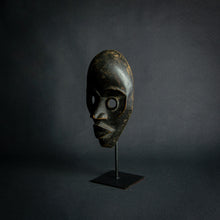 Load image into Gallery viewer, an mask from West Africa featuring round eyes and a serene expression, mounted on a stand against a dark background.