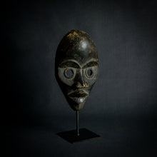 Load image into Gallery viewer, an mask from West Africa featuring round eyes and a serene expression, mounted on a stand against a dark background.