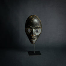 Load image into Gallery viewer, an mask from West Africa featuring round eyes and a serene expression, mounted on a stand against a dark background.