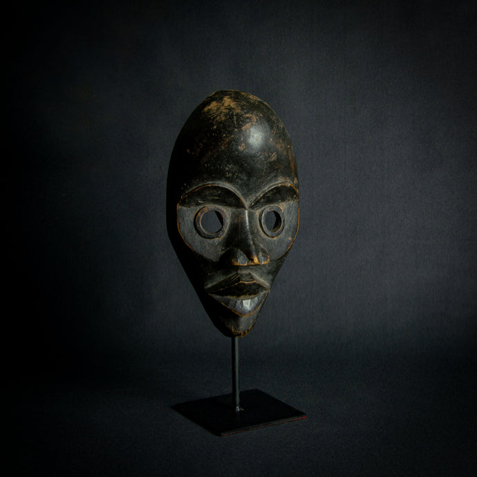 an mask from West Africa featuring round eyes and a serene expression, mounted on a stand against a dark background.